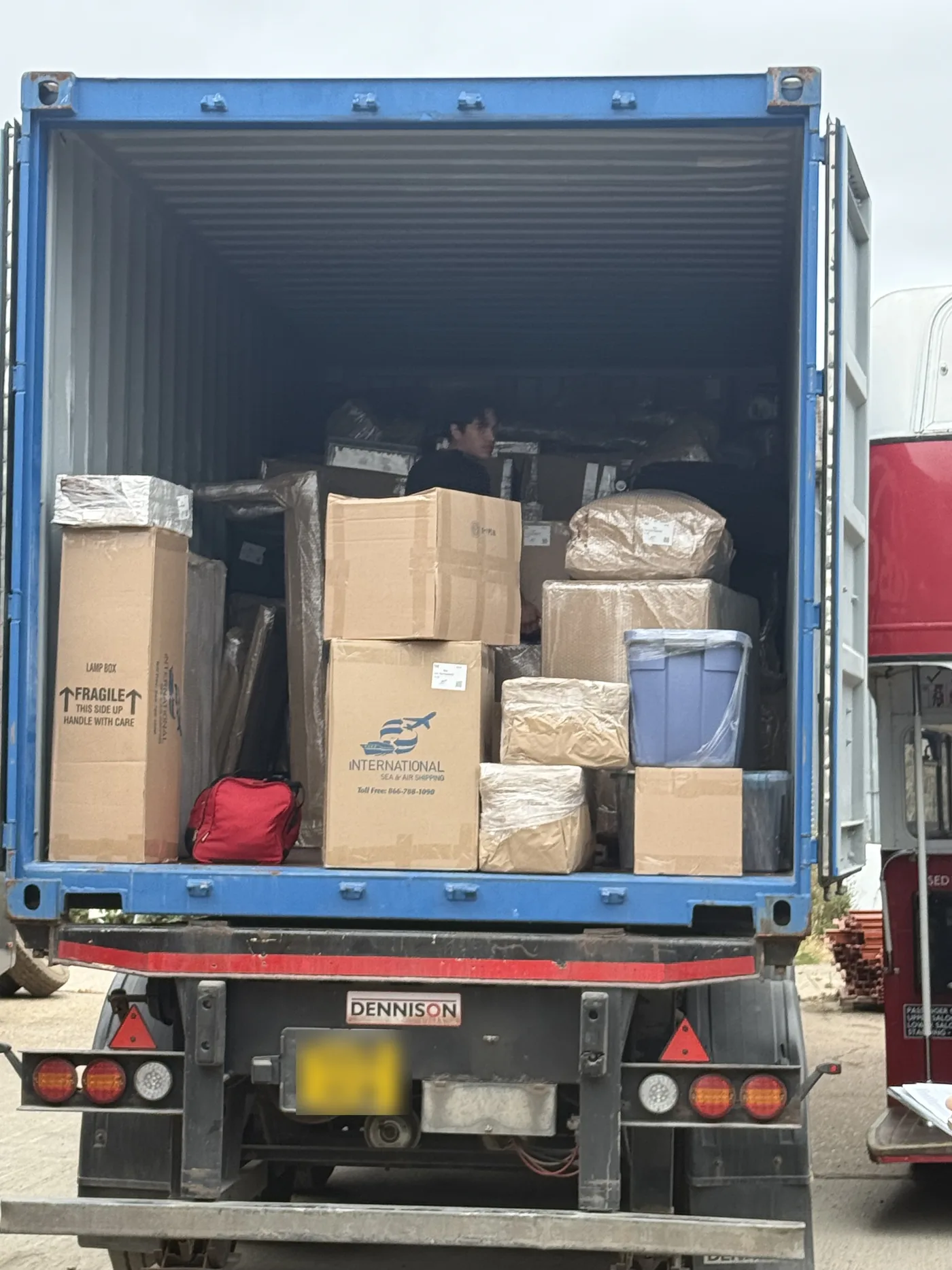Open shipping container packed with cartons, wrapped furniture and a lamp box, being unloaded